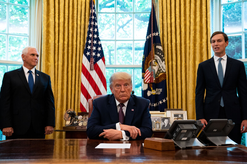 Man sits behind a desk, two men stand behind him