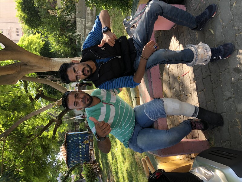 Two men, both with bandages on their legs, sit on a park bench giving the thumbs-up.