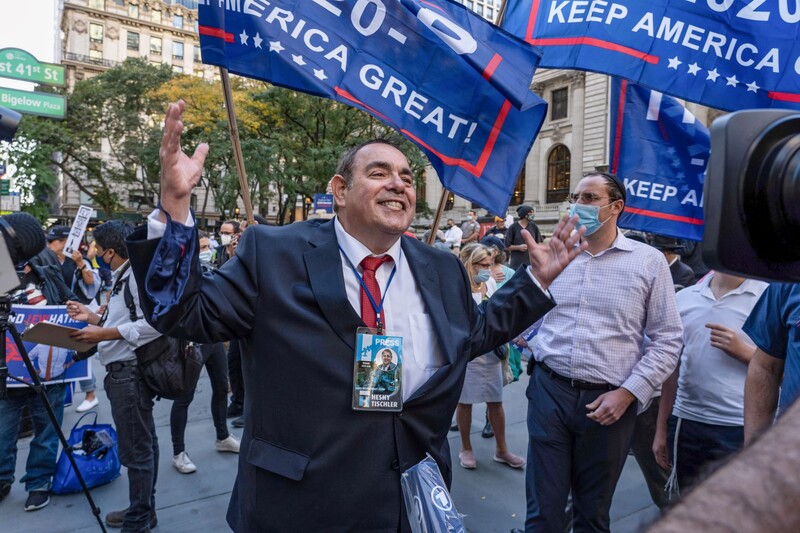 Man speaks with upraised arms with Trump flags behind him