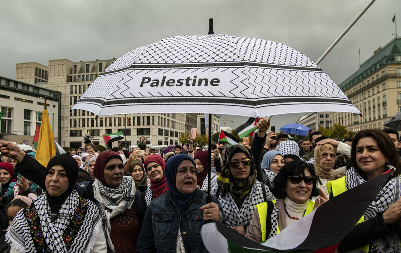 A group of women stand beneath a black-and-white umbrella emblazoned with the word 'Palestine'. 