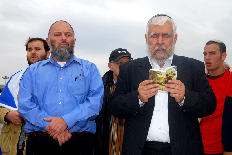 Standing men look ahead as one reads from a book