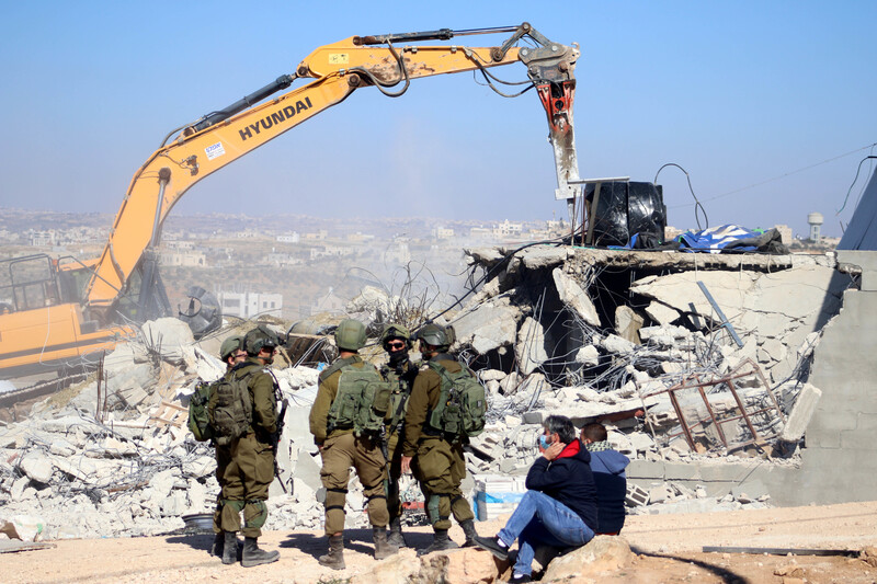 Heavily armed soldiers watch Hyundai bulldozer demolish a home while two men observe nearby