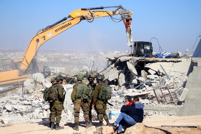 Heavily armed soldiers watch Hyundai bulldozer demolish a home while two men observe nearby 
