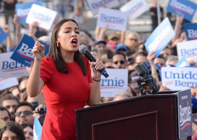 Woman speaks at podium in front of large crowd carrying signs 