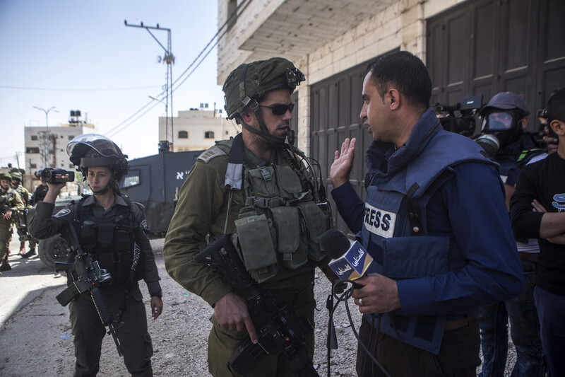 An armed Israeli soldier confronts a man wearing a press flak jacket while holding a microphone