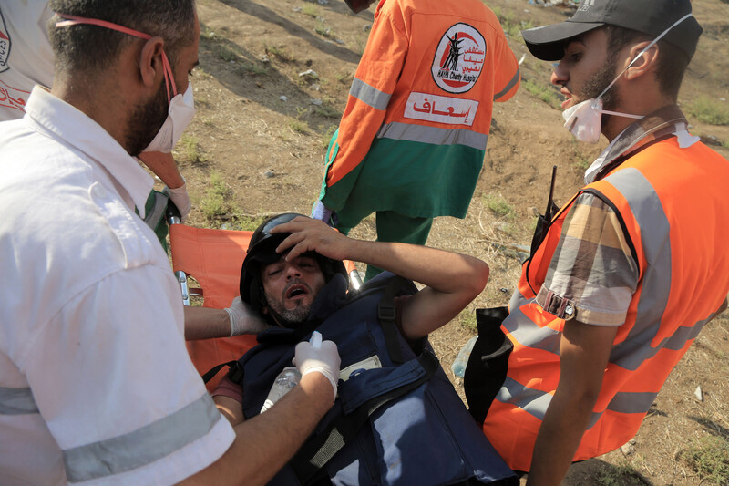 A wounded man wearing a press helmet and flak jacket is carried on a stretcher