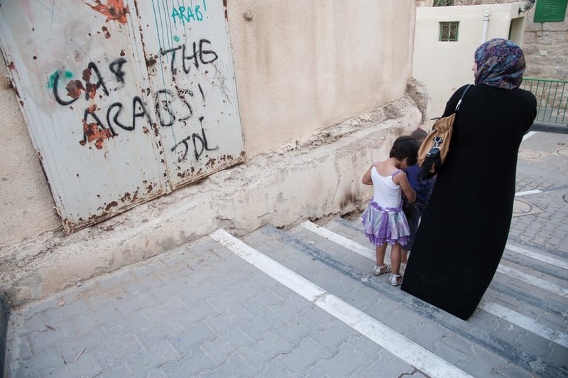 Woman and children walk past wall spray painted with graffiti reading Death to the Arabs!