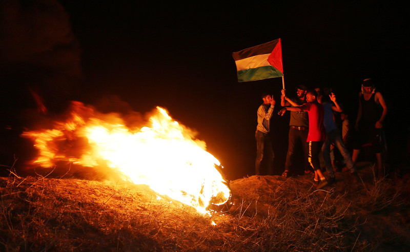 A small group of people stand next to a fire holding a Palestinian flag in the dark