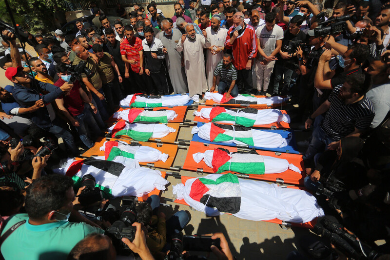 A crowd of men stand around ten bodies shrouded in Palestinian flags