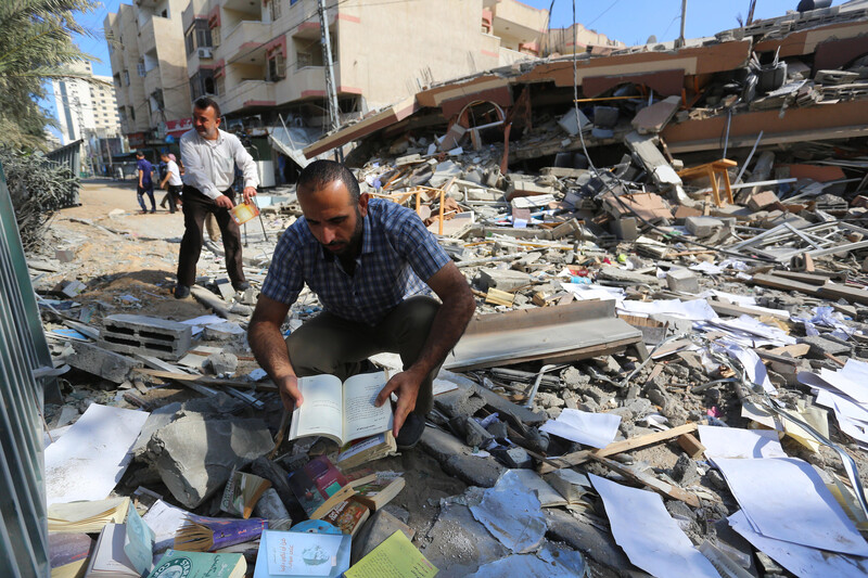 A man kneeling on the ground holds an open book in his hands amid rubble from a collapsed building