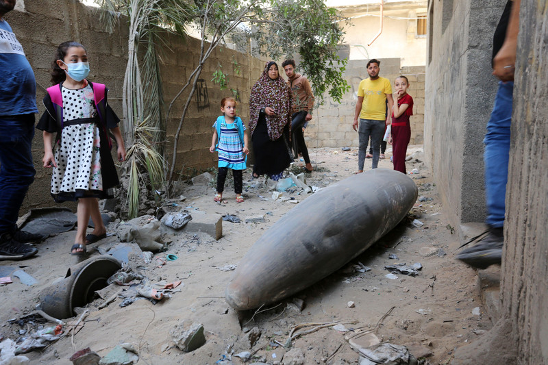 Men, women and children stand near large missile in narrow alley