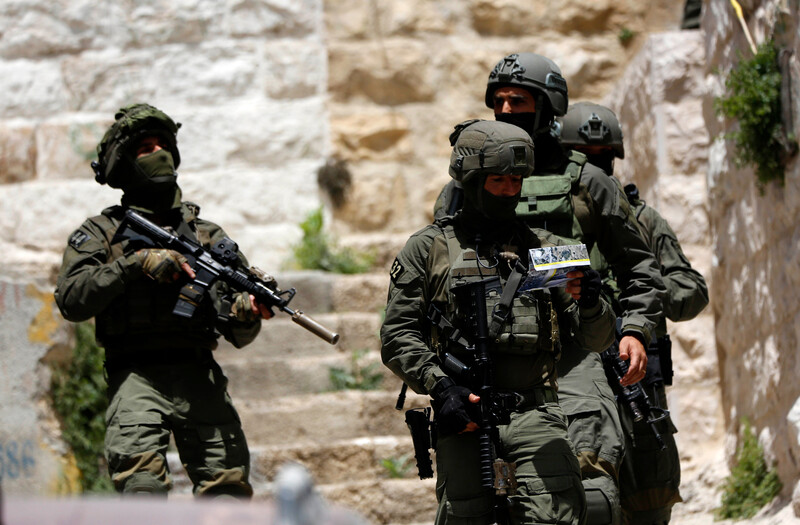 Four heavily armed Israeli soldiers stand near stone building 