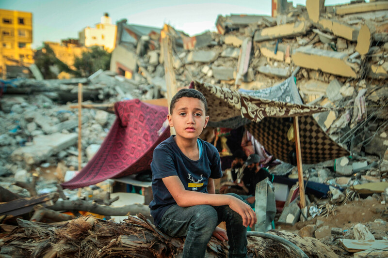 Boy sits on rubble in front of makeshift tent amid destroyed buildings