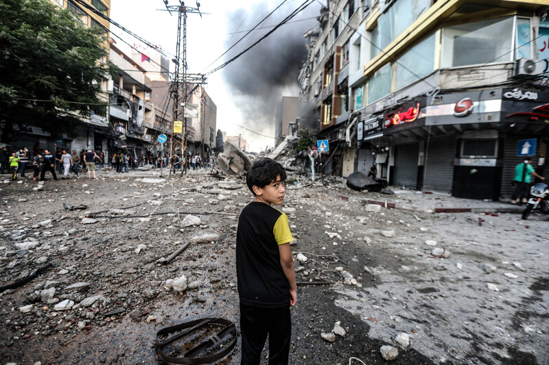 A boy stands in debris-strewn street with smoke on the horizon