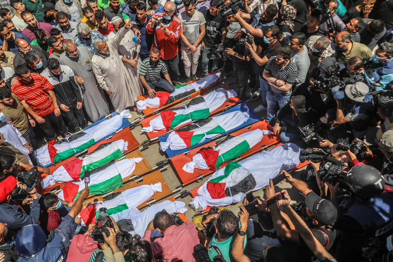 People stand around 10 bodies on the floor wrapped in Palestinian flags 