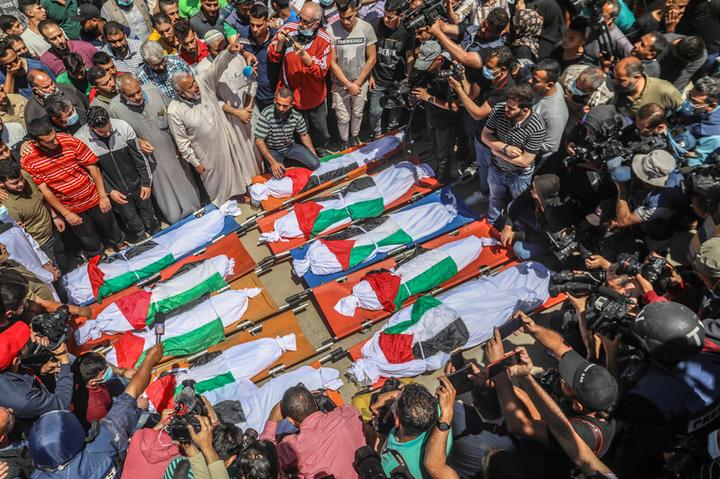 People stand around 10 bodies on the floor wrapped in Palestinian flags