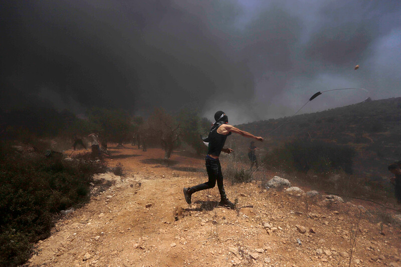 Standing in a smoke-filled field, a masked protester hurls a stone with a slingshot  