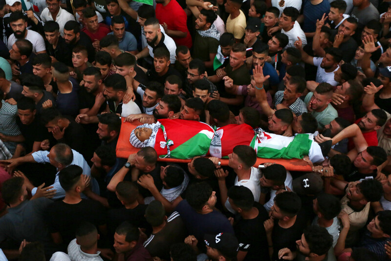 A large crowd of mourners carry a body wrapped in a Palestinian flag on a stretcher 