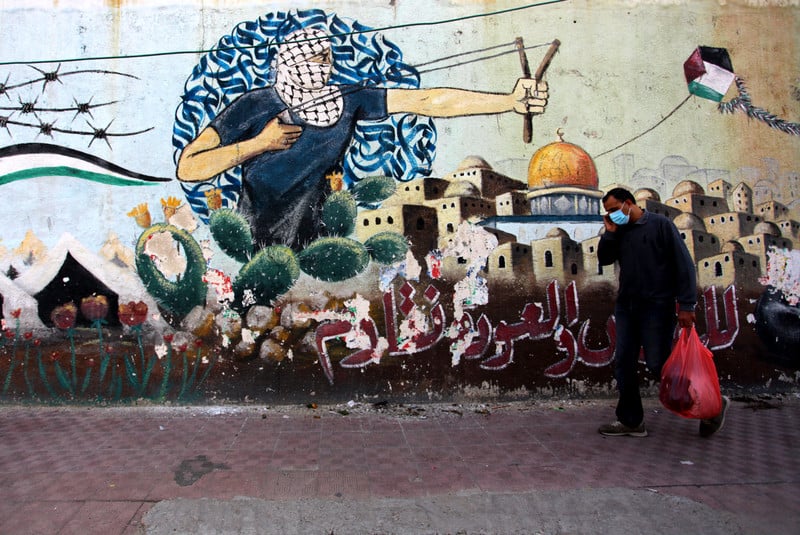 A man with a shopping bag walks past graffiti depicting the Dome of the Rock and keffiyeh-clad youth wielding a slingshot 