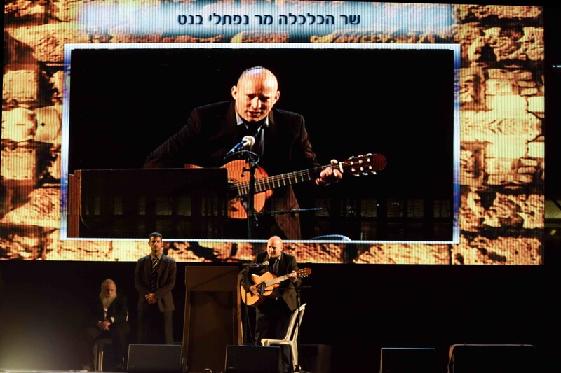 A giant screen shows one man playing a guitar as three people stand on stage beneath