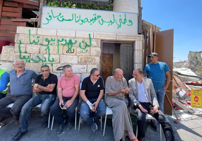 Seven men sit in front of a graffitied building, near rubble