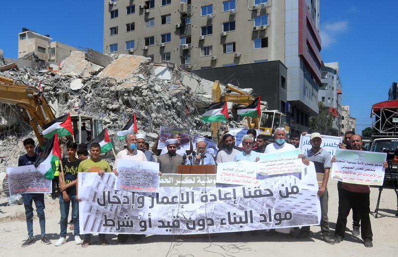 People hold banners and flags before a podium against a background of rubble 