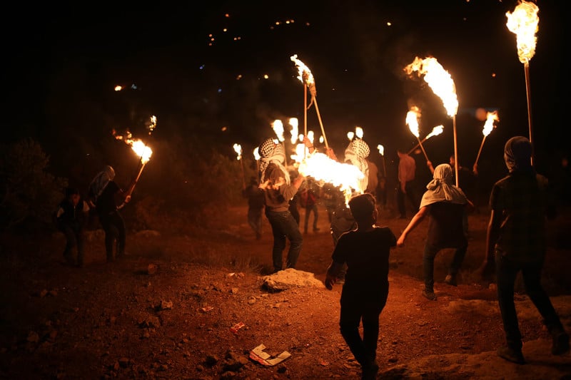 A group of men with lit torches walk across a field in the night