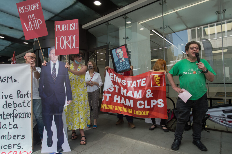 Man speaks into microphone as people around him hold signs and banners