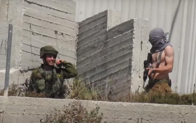 Smiling man in military uniform gestures towards man with shirt pulled over his head carrying rifle