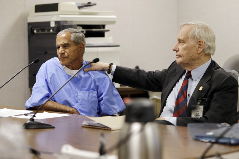 Man in suit places hand on shoulder of man in prison uniform seated next to him