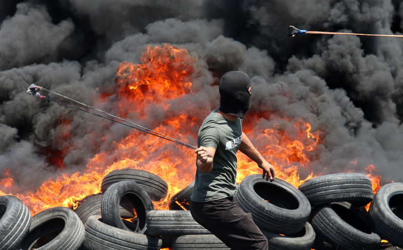A man wields a slingshot in front of burning tires