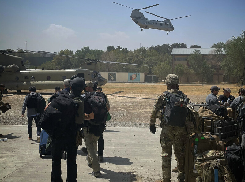 A helicopter lifts off as people holding baggage stand nearby