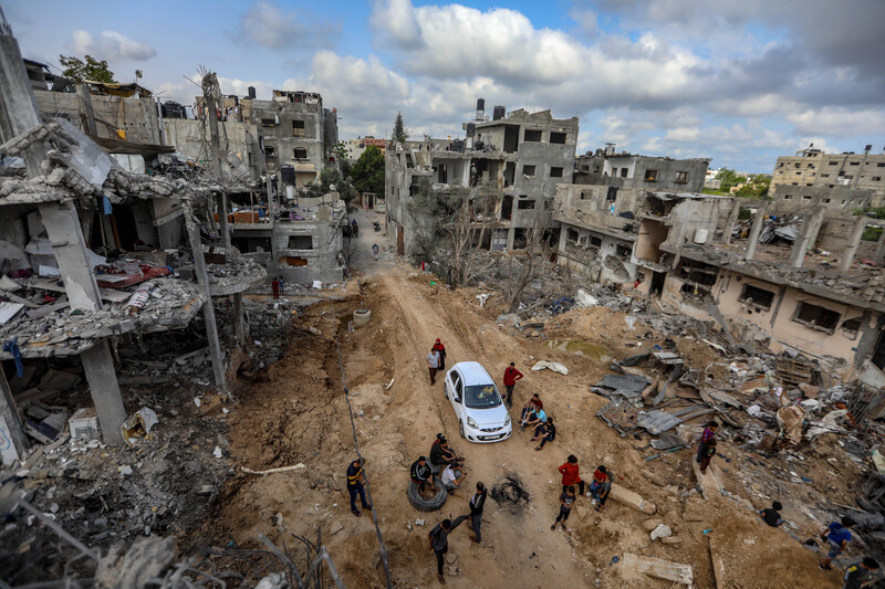 Aerial view of a car and people on a dirt-covered road surrounded by bombed-out buildings