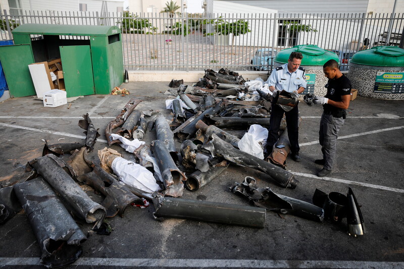 Two men handle pieces of rockets while standing next to dozens of metal cylinders in parking lot 