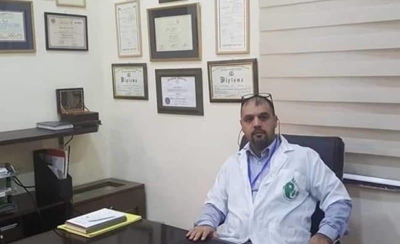 Man in medical uniform sits at desk with framed diplomas on wall behind him