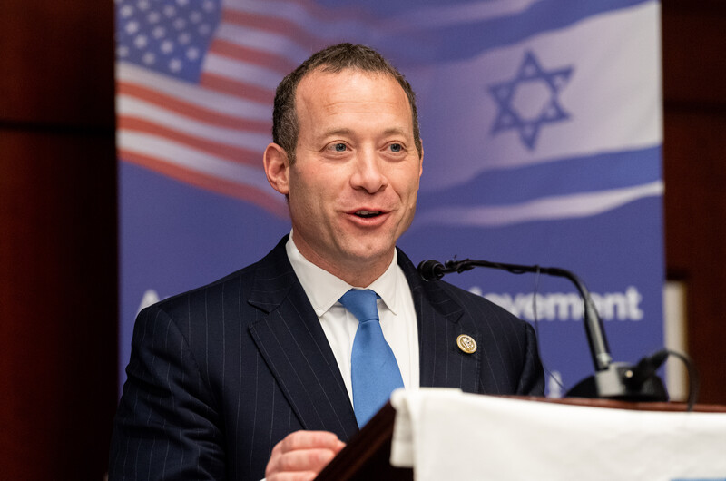 A man in a suit stands in front of US and Israeli flags