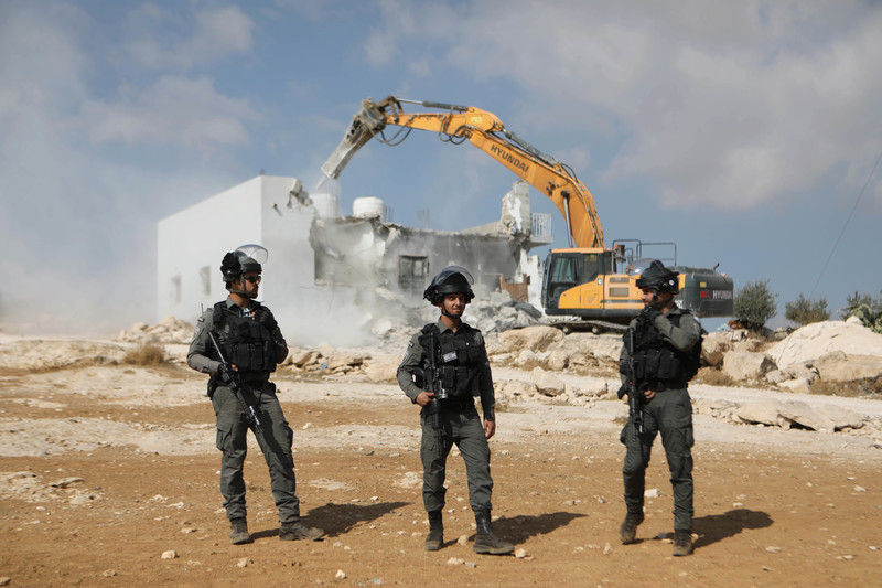 Three armed men stand in front of half destroyed house and construction machine