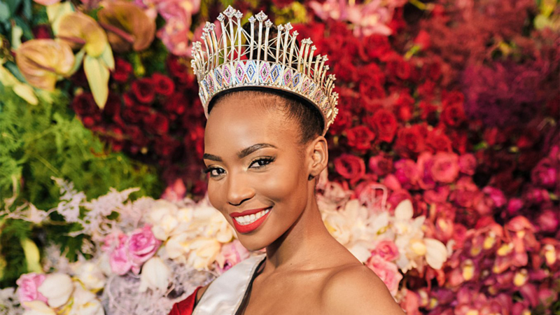 A woman wearing a crown and sash against a backdrop of flowers 