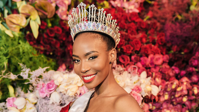 A woman wearing a crown and sash against a backdrop of flowers