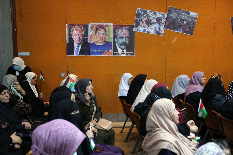 A group of people sit beneath three posters depicting British PM Boris Johnson, British minister Pirit Patel and Arnold Balfour, who penned the Balfour declaration, giving away Palestine