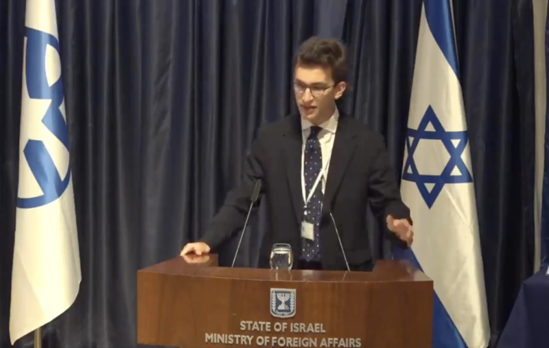 A young man speaks from a lecturn surrounded by Israeli flags