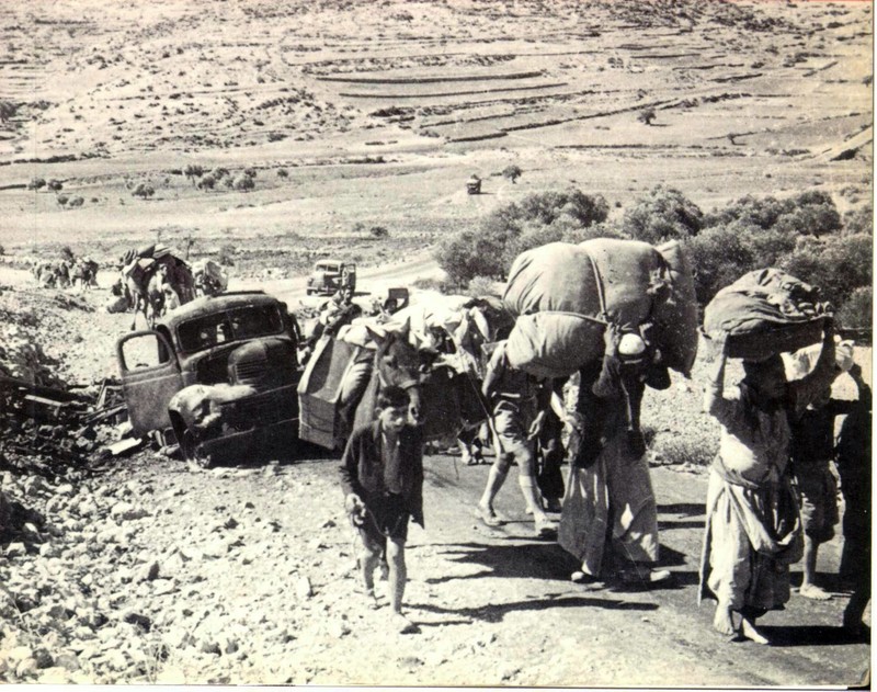A black and white photo shows people on foot carrying heavy burdens passing a broken down vehicle