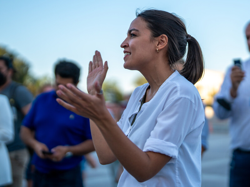 Woman speaks at microphone with onlookers shown behind her