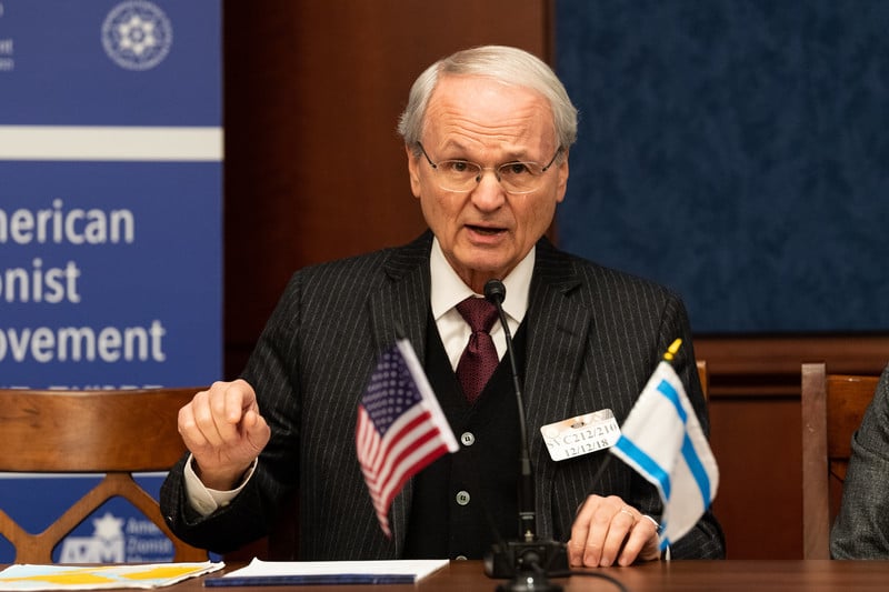 Photo of Morton Klein gesticulating with microphone and small American and Israeli flags in front of him