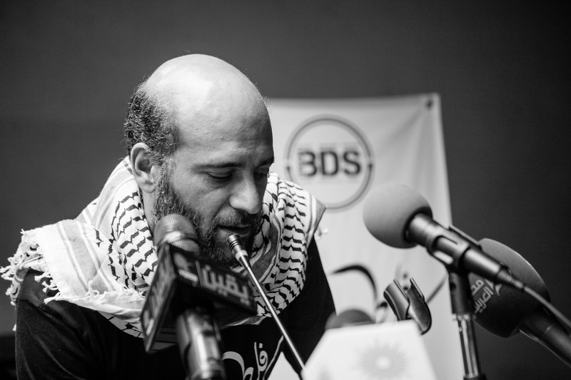 A man wearing a checkered scarf speaks to multiple microphones