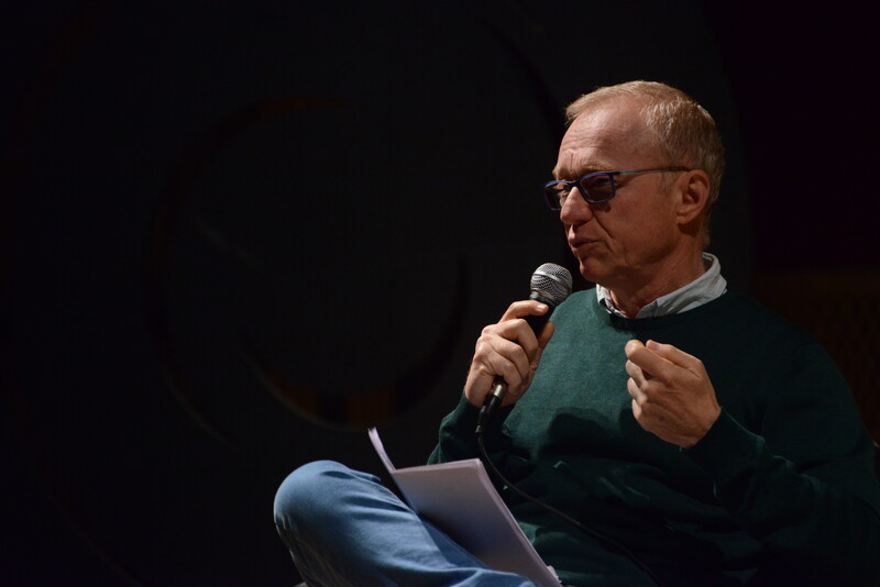 A man in glasses speaks into a microphone with a paper in his lap 