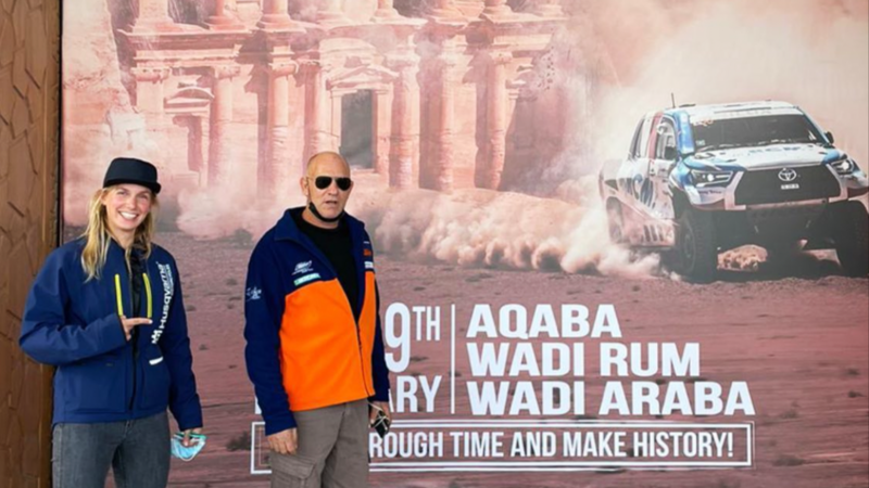 A man and a woman pose in front of a large poster featuring a racing car 