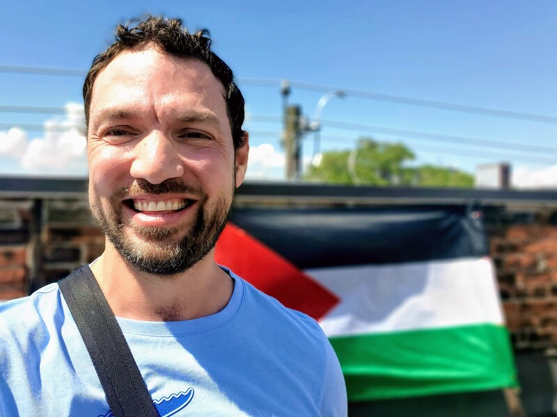 A man smiles in front of a Palestinian flag