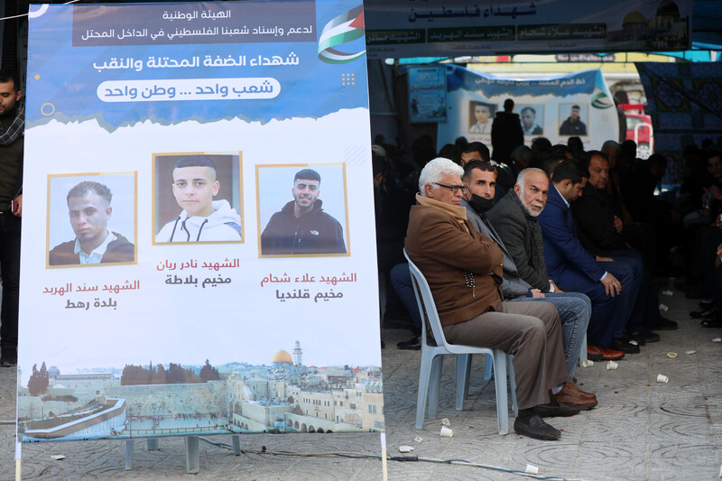 Men sit on plastic chairs under a large tent next to a banner featuring pictures of three men 