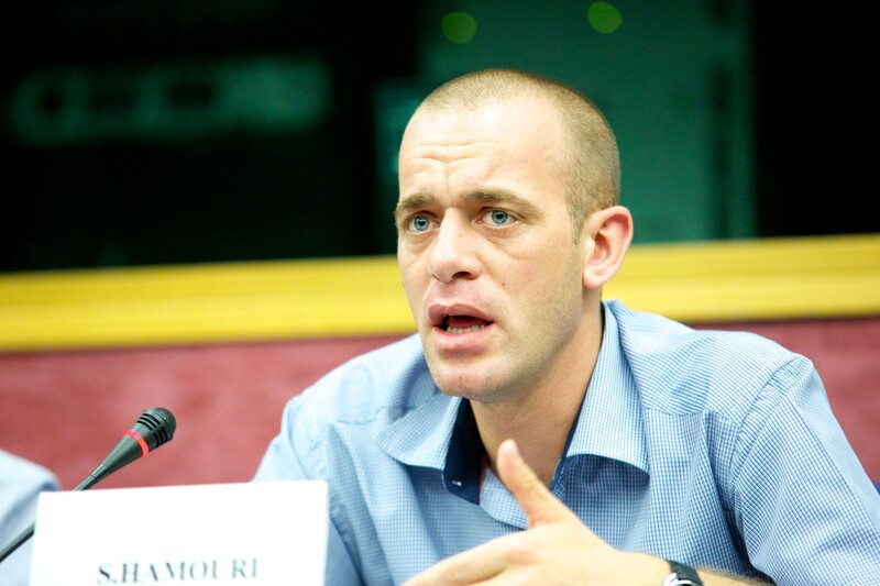 A man talks in front of a microphone and a name card 